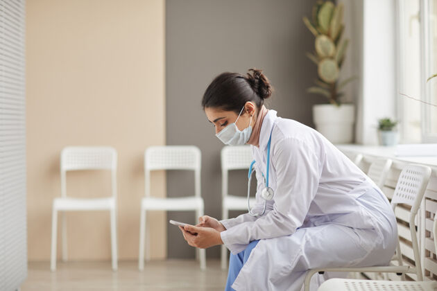 成人穿着白大褂 戴着口罩的女医生坐在走廊边用手机打字看互联网室内
