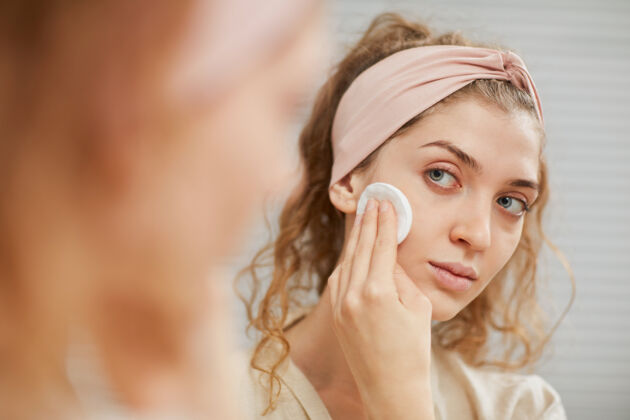 皮肤护理长卷发的年轻女子站在镜子前用海绵擦脸特写美容产品肤色