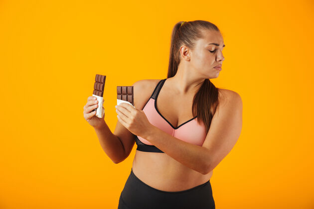 女人一个穿着运动服的心烦意乱的超重年轻女子的肖像 孤立地站在黄色的墙上 手里拿着巧克力棒饮食站身体