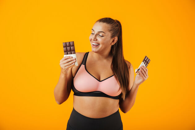 女运动员一个穿着运动服的快乐超重的年轻女子孤立地站在黄色的墙上 吃着巧克力墙壁高兴食物