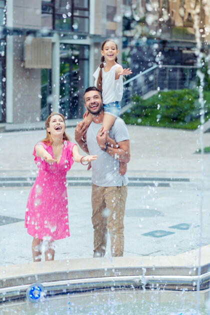男人水的清新站在喷泉旁看着水滴的快乐的一家人水女儿家庭