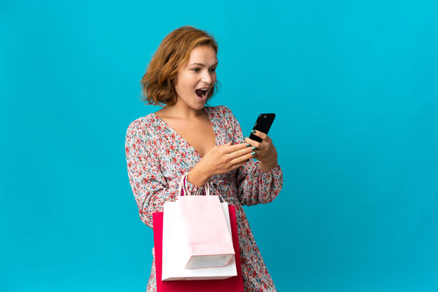 包一位格鲁吉亚年轻女子拿着购物袋孤零零地站在蓝色的墙上 手里拿着购物袋 用手机给一位朋友留言购物者礼品商业
