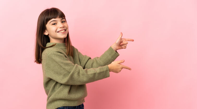 小小女孩孤立地站在粉红色的墙上 手指着侧面 展示着一个产品介绍手指产品