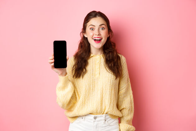 情绪科技和网上购物迷人的女学生看起来很兴奋 显示空的智能手机屏幕和微笑 站在粉红色的墙壁在线兴奋魅力