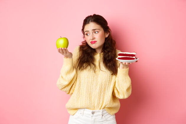 情绪一个年轻的女人抱着一块美味的蛋糕 面带悲伤和失望地看着青苹果 想吃甜点 站在粉红色的墙上女人苹果兴奋