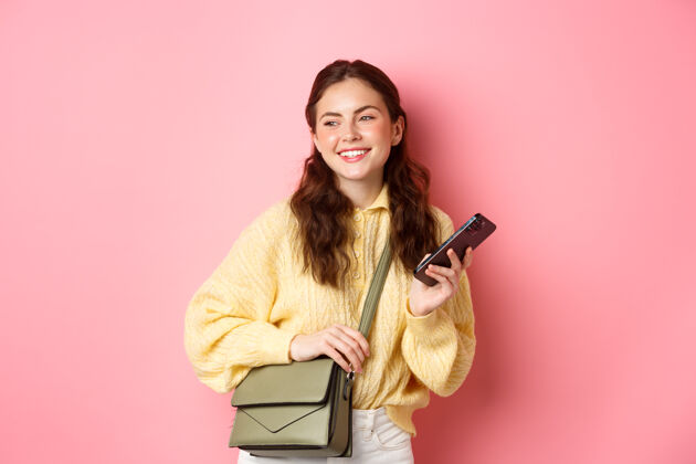 黑发时髦的现代年轻女子拿着智能手机和钱包 站在粉红色的墙上 开心地微笑着向旁边看微笑微笑候选人