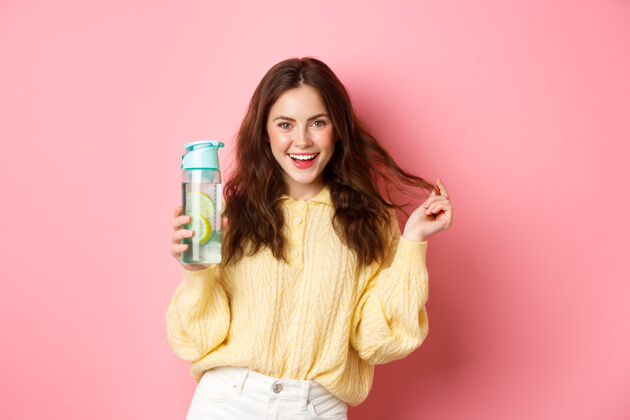 女性性感迷人的女人自信地看着镜头 拿着装有柠檬水的健康饮料的私人瓶子 站在粉色的墙上积极兴奋女孩