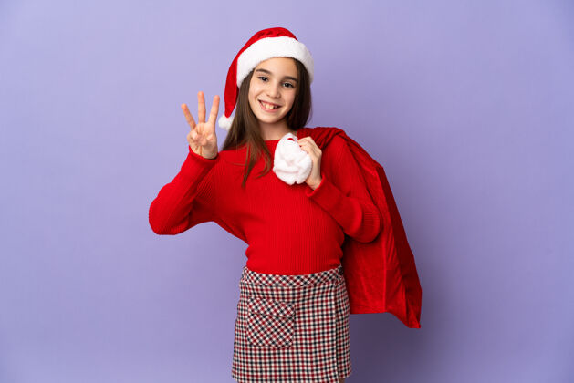 3戴着帽子和圣诞袋的小女孩被隔离在紫色的墙上 快乐地用手指数着三个33童年