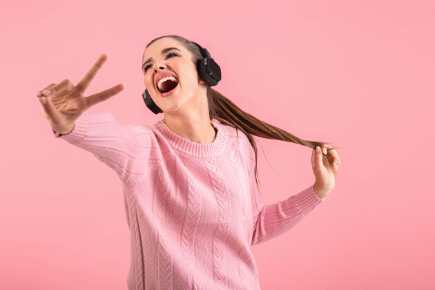 立体声年轻迷人的女人戴着无线耳机听音乐穿着粉色毛衣微笑着快乐积极的心情摆在粉色背景上配件时髦音乐