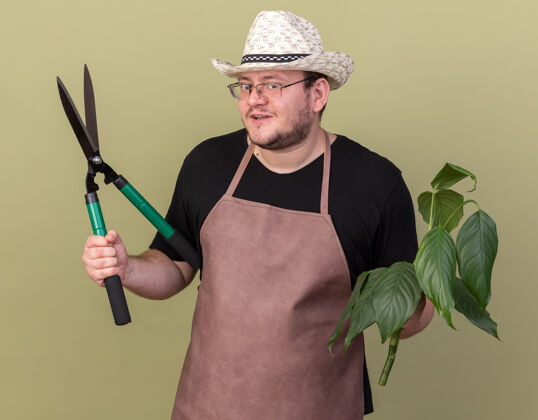 男高兴的年轻男园丁戴着园艺帽拿着修剪器隔离在橄榄绿墙上的植物感情站脸