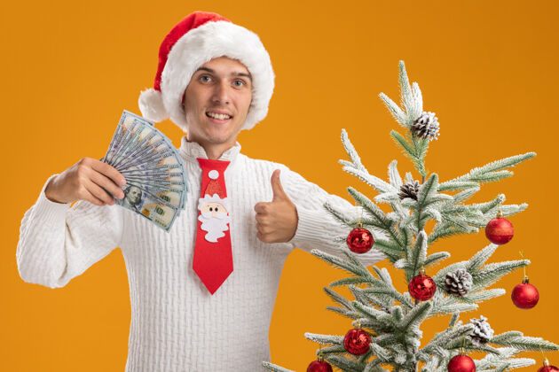 近微笑着的年轻帅哥戴着圣诞帽 打着圣诞老人的领带 站在装饰好的圣诞树旁 手里拿着钱 神情孤零零地竖起大拇指站在橙色的墙上帅哥圣诞老人帽子