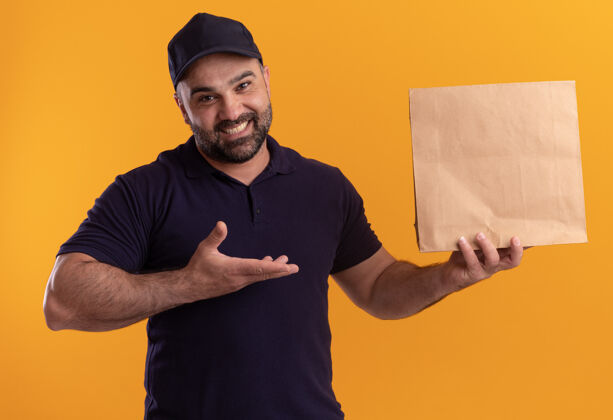 公民身着制服 戴着帽子 面带微笑的中年送货员指着隔离在黄色墙上的纸食品包装感觉表情姿势