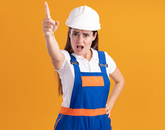人穿着制服的年轻建筑女严格地指着镜头 把手放在隔离在橙色墙上的臀部上制服点表情