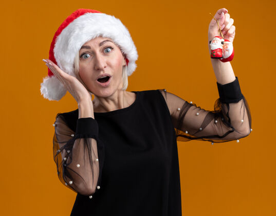 圣诞节惊讶的中年金发女人戴着圣诞帽看着举起圣诞老人的圣诞饰品保持手靠近头部隔离在橙色的墙上保持提高女人