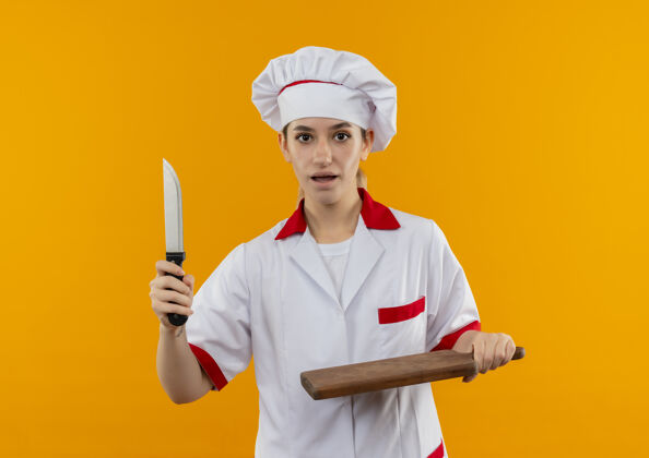 制服年轻漂亮的厨师穿着厨师制服拿着刀和砧板孤立在橙色的墙上漂亮印象刀