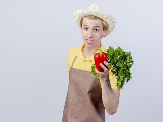 帽子年轻的园丁穿着连体衣 戴着帽子 手里拿着红甜椒和新鲜的草药 脸上带着微笑红色年轻站着