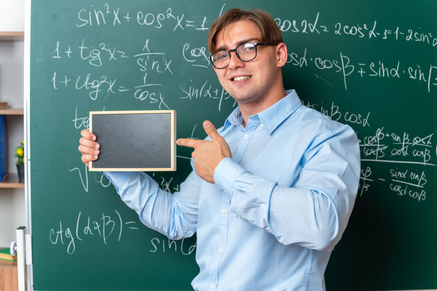 教室年轻的男老师戴着眼镜 拿着小黑板 用食指指着黑板 微笑着自信地站在教室里数学公式的黑板旁年轻指向公式