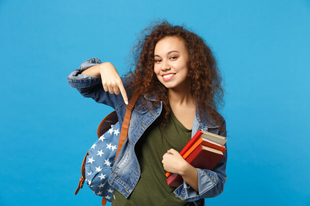 手指穿着牛仔服 背着书包的年轻学生把书孤立地放在蓝色的墙上非洲智能地方