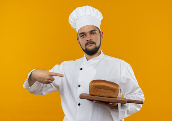 拿着身着厨师制服的自信的年轻男厨师手拿着并指着橘色墙上挂着面包的砧板制服自信男性