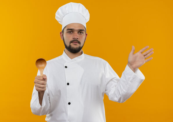 显示自信的年轻男厨师穿着厨师制服 拿着勺子 在橘色的墙上展示着孤立的空手信心手烹饪