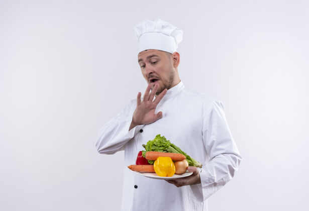 印象年轻帅气的厨师穿着厨师制服 手里拿着盛有蔬菜的盘子 手放在上面 放在隔离的白色墙壁上 留着复印空间烹饪持有制服