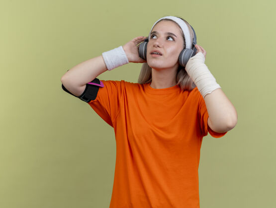 运动惊讶的年轻白人运动女孩戴着耳机戴着护腕戴着头带和手机臂带抬头看绿色背带袖带