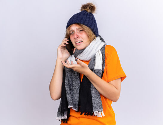 痛苦不健康的年轻女子穿着橙色t恤 脖子上围着暖和的围巾 戴着帽子 感觉很糟糕 患了流感 在手机上说 表情悲伤 站在白色的墙壁上担心女人围巾表情