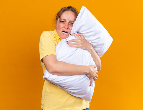 站立穿着黄色衬衫的不健康的女人感觉不舒服 患流感 站在橙色的墙上抱枕受凉不适女人枕头
