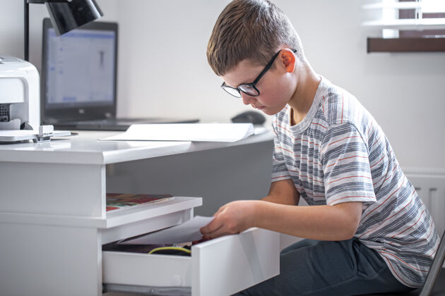 在线学校戴眼镜的小男孩坐在笔记本电脑前的桌子旁 在抽屉里找东西灯学生小