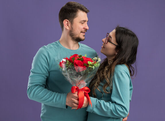 随意年轻漂亮的一对穿着蓝色休闲服的男女捧着一束玫瑰看着彼此相爱的幸福一起站在紫色的墙上玫瑰一起情侣