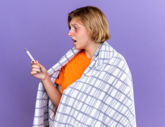 流感不健康的年轻女子裹着温暖的毯子 感觉不舒服 患流感 发烧 用温度计测量体温 站在紫色的墙上 看起来很担心使用担心包裹