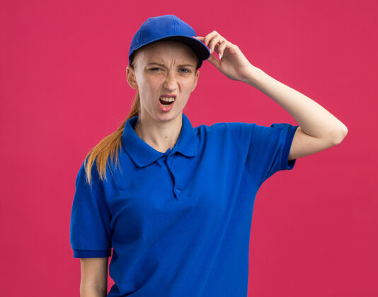 年轻穿着蓝色制服 戴着帽子的年轻送货员 站在粉红色的墙上 误把手放在头上 弄糊涂了手困惑女孩