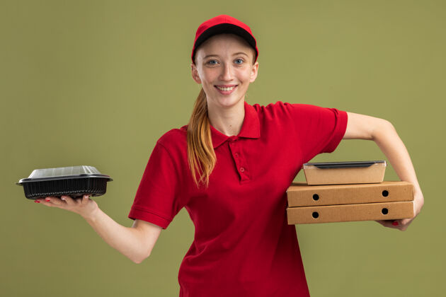 帽子身穿红色制服 头戴鸭舌帽 手持比萨饼盒和食品包的年轻送货女孩 自信地微笑着越过绿墙女孩制服送货