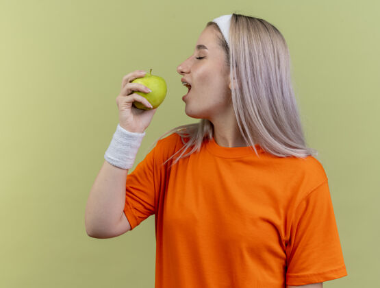 咬高兴的年轻白人运动女孩戴牙套戴头带和手腕假装咬苹果苹果橄榄假装