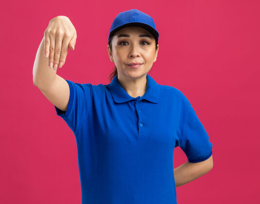手身穿蓝色制服 头戴帽子的年轻女送货员自信地指着站在粉红色墙上的手立场表情年轻