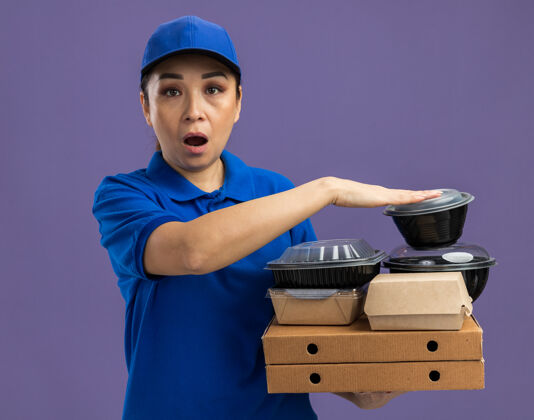 制服身穿蓝色制服 头戴鸭舌帽 手持比萨饼盒和食品包的年轻送货员站在紫色的墙上 既惊讶又惊讶帽子拿着站着
