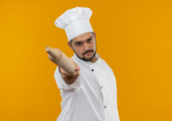 男性自信的年轻男厨师身着厨师制服 将擀面杖伸向孤立的橙色墙壁 留有复制空间别针伸展滚动