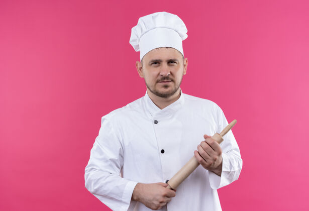 帅气自信的年轻帅哥厨师穿着厨师制服拿着擀面杖孤立地站在粉红色的墙上持枪滚烹饪