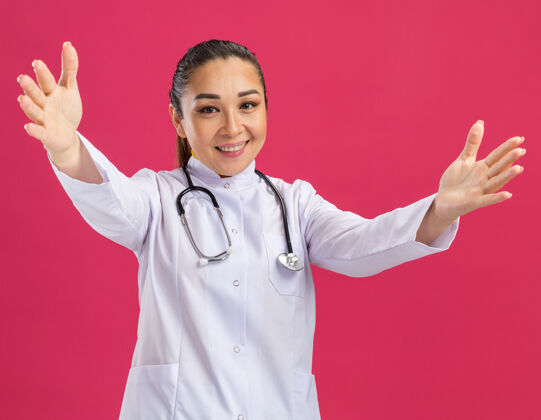 手快乐的年轻女医生 穿着白色药衣 脖子上戴着听诊器 做着欢迎的手势 张开双手站在粉红色的墙上药年轻人做