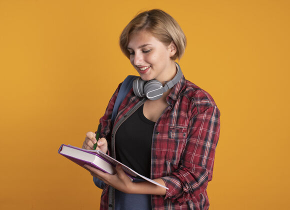 写作微笑的年轻斯拉夫女学生戴着耳机 背着背包 用笔在笔记本上写字复制橙色年轻人