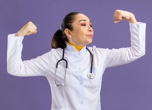 医生身穿白色药衣 脖子上戴着听诊器的年轻女医生站在紫色的墙上 举起拳头 看上去很自信周围外套举起来