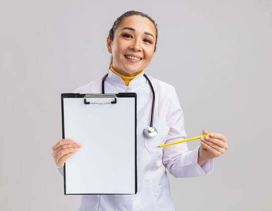 外套年轻的女医生穿着白色的医疗外套 脖子上戴着听诊器 拿着空白页的剪贴板 站在白色的墙上微笑着要签名医生翻页铅笔