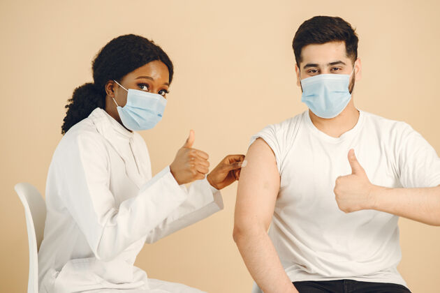 批准隔离 米色背景非洲女医务工作者和戴着口罩的男子竖起大拇指接种疫苗对抗柯维德流行病疾病大流行