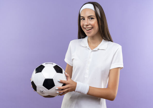 球快乐漂亮的运动女孩戴着头带和手环拿着足球隔离在紫色的墙上穿漂亮腕带