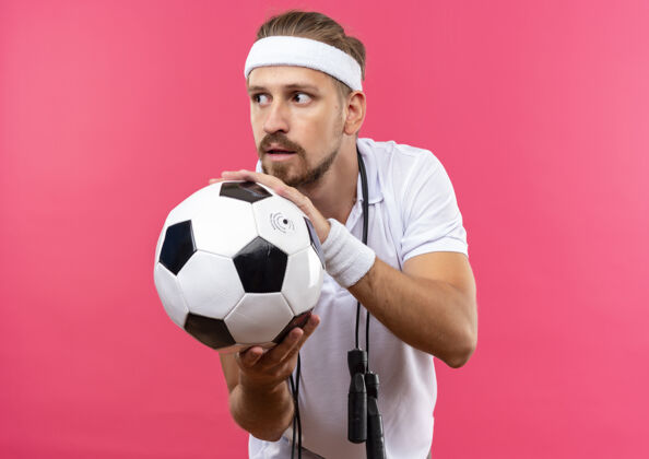 腕带令人印象深刻的年轻英俊的运动男子戴着头带和手镯拿着足球看一边跳绳围着脖子隔离在粉红色的墙上脖子壁板穿
