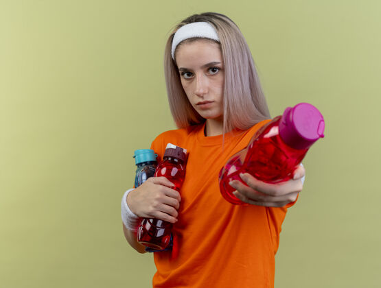 复制自信的年轻白人运动女孩戴着背带 戴着头带和腕带 拿着水瓶看着镜头瓶子年轻绿色