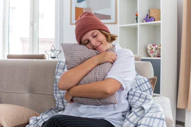 女人不健康的年轻女子戴着暖和的帽子 裹着毛毯 患感冒和流感 抱着枕头 闭着眼睛微笑 坐在明亮的客厅沙发上年轻痛苦毯子
