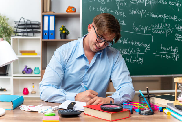 老师戴眼镜的年轻男老师坐在课桌旁 在教室的黑板前写着书和笔记男写字前面