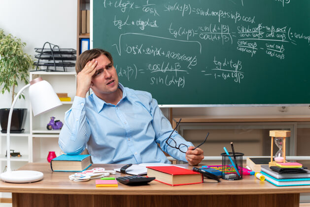 数学年轻的男老师戴着眼镜 看起来很累 工作过度 摸着头坐在课桌旁 教室的黑板前放着书和笔记眼镜超负荷工作笔记