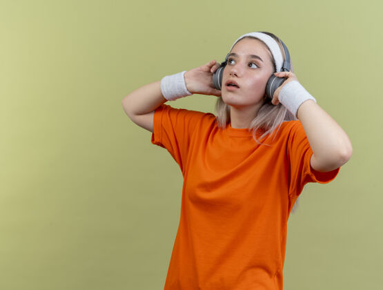 腕带令人印象深刻的年轻白人运动女孩戴着背带耳机戴着头带和腕带看一边复制运动壁板